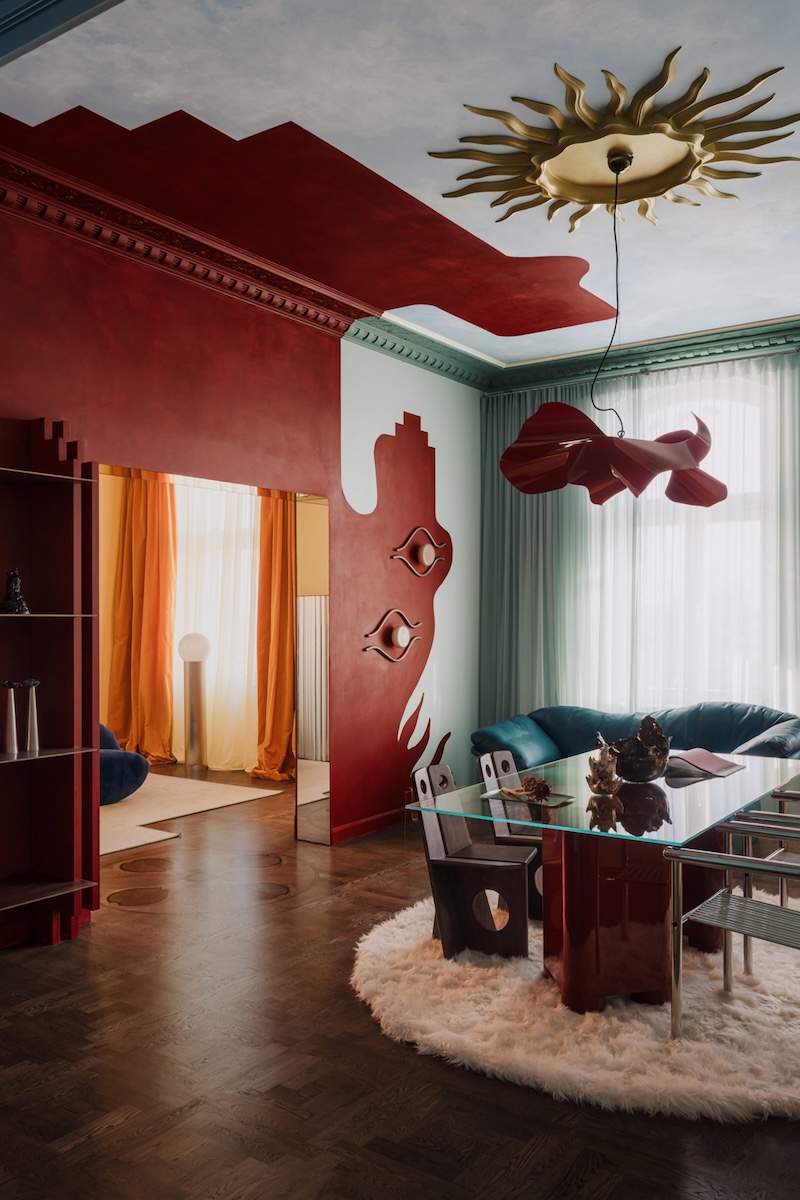 Interior of the Two Suns apartment by COI Studio with a striking red wall, historic stucco and contrasting design elements including a glass table and coloured curtains.