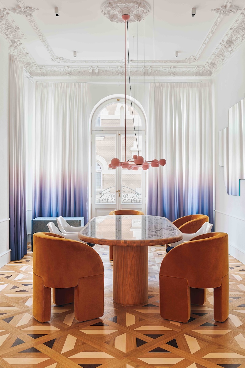 Dining room in a historic interior with an ornamental ceiling, gradient curtains in shades of pink and blue, a wooden table and a designer pendant light