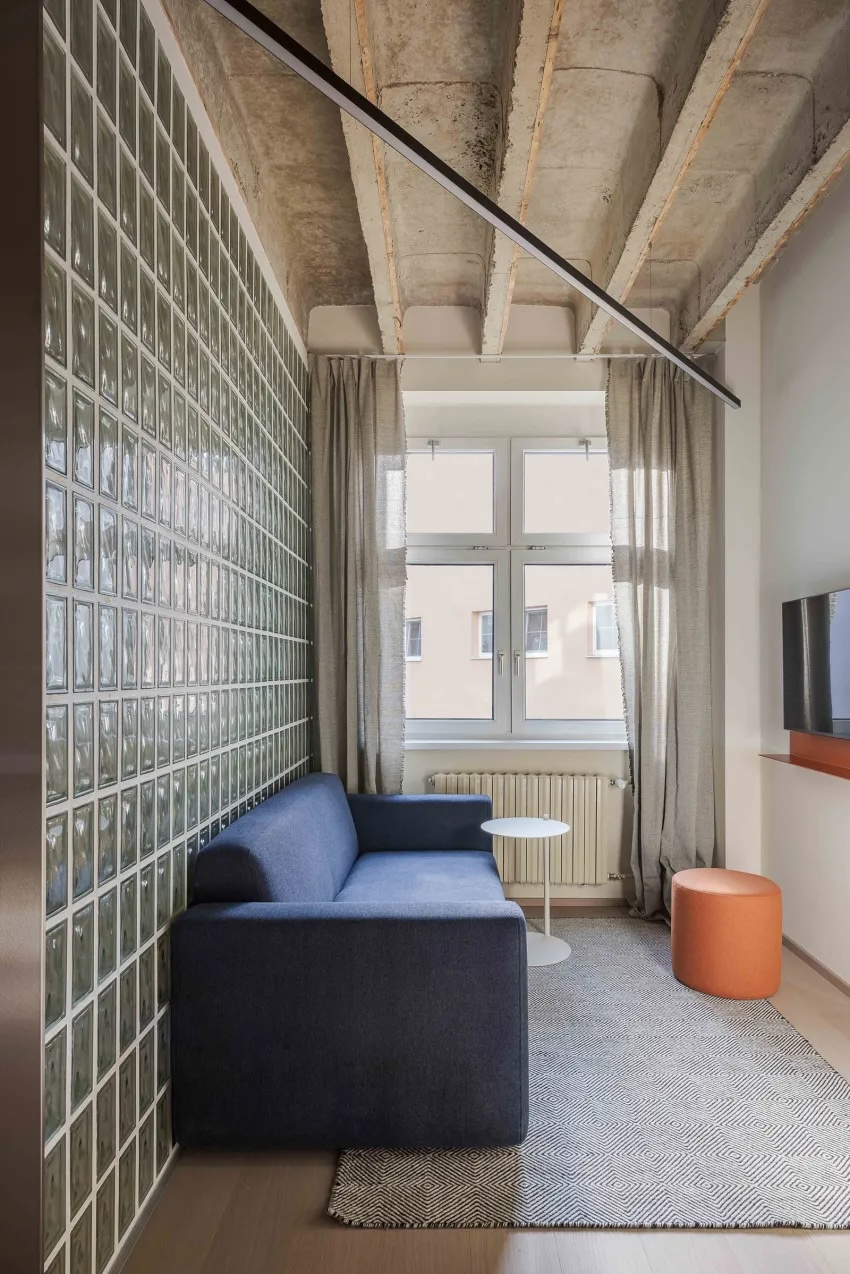 Moderní interiér od BEEF Architekti s modrou pohovkou, stěnou ze skleněných tvárnic, pohledovým betonovým stropem a minimalistickým nábytkem v neutrálních tónech.