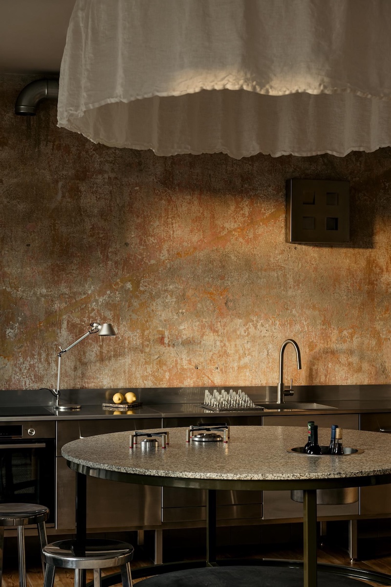 Kitchen by Wierciński Studio with a patinated textured wall, stainless steel counter, stone island and minimalist lighting